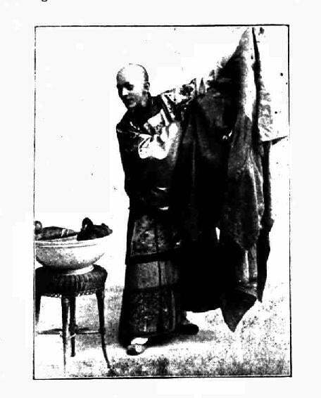 Very old black and white image of a Chinese magician in traditional dress. His left arm is outstretched an dhe is looking down at a small table.
