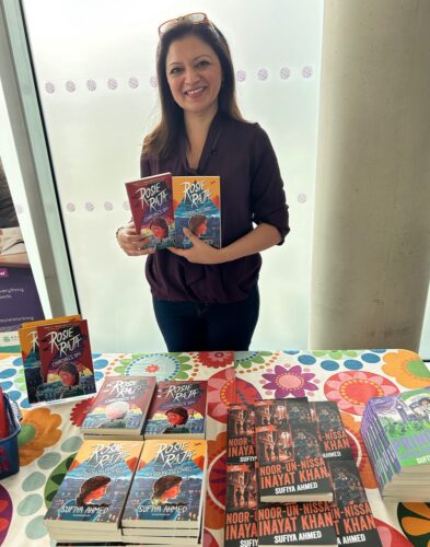 Sufiya Ahmed holding copies of her Rosie Raja series books which are also laid out on a table in front of her
