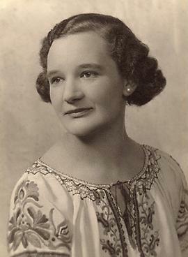 A woman with short 1920s waved hair looking into the distance. She is wearing a white blouse with flower embroidery and white pearl earrings.