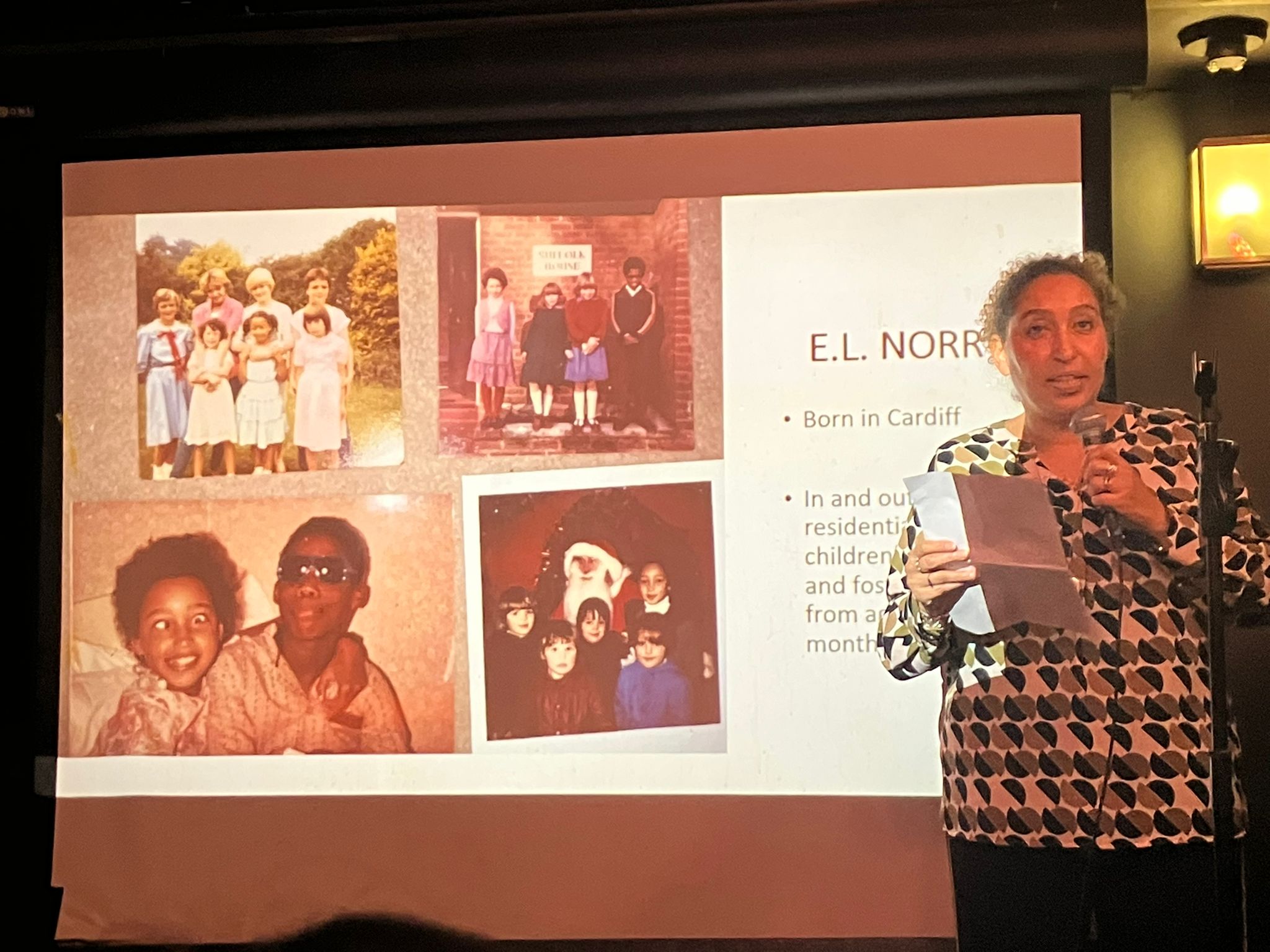 E.L.Norry presenting in front of a Powerpoint presentation slide showing pictures of her childhood