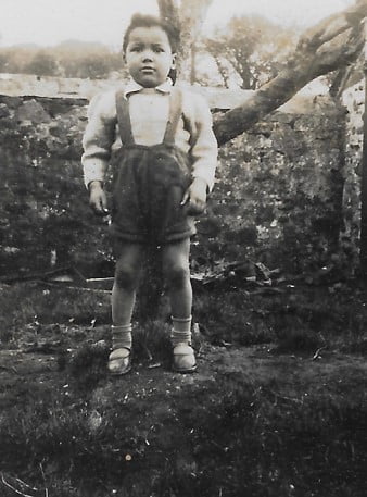 A black and white photo of a young mixed race boy standing in a typically English back garden circa 1940s.