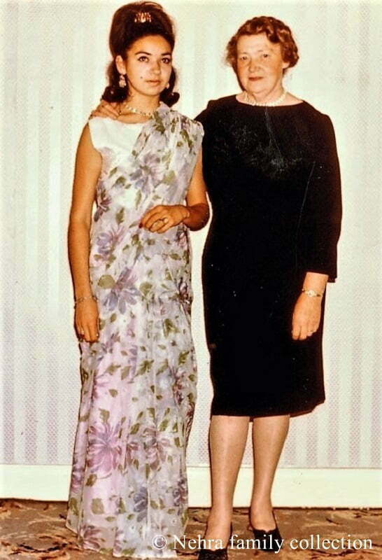 A young mixed race woman poses during the late 1960s at home in a sari next to her white middle aged white mother, dressed in black.