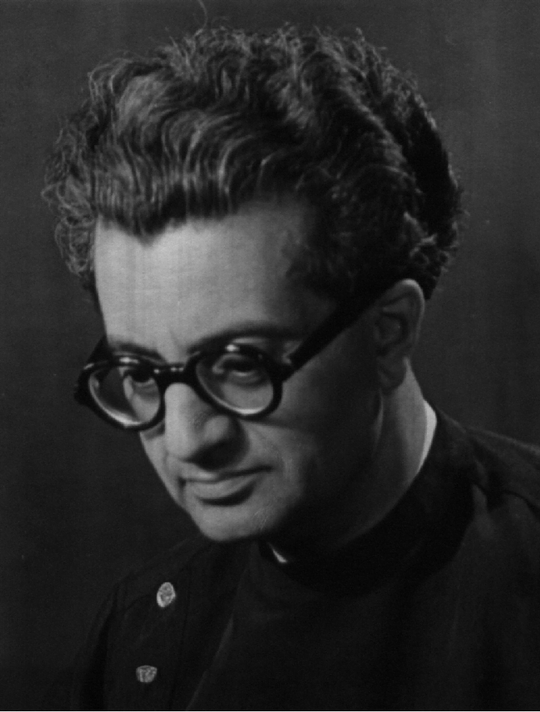 Photograph a man with dark wavy hair looking down. He is wearing a black jacket with round buttons and thick black rimmed glasses.