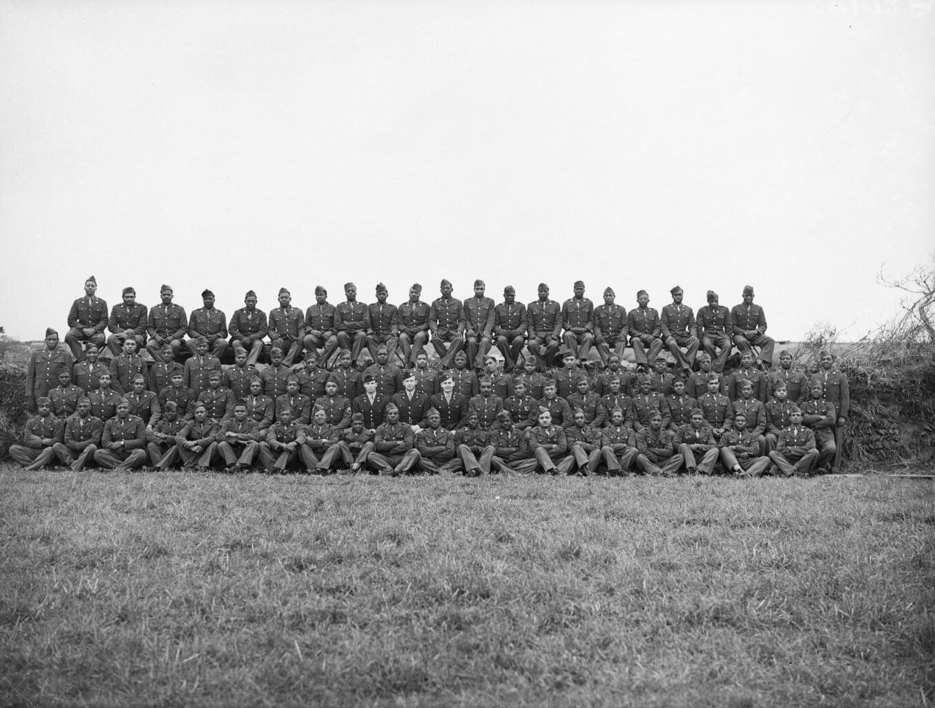 Large group of Black GIs lined up in rows for an official photograph in an English field. White commanding officers are in the centre of the rows.