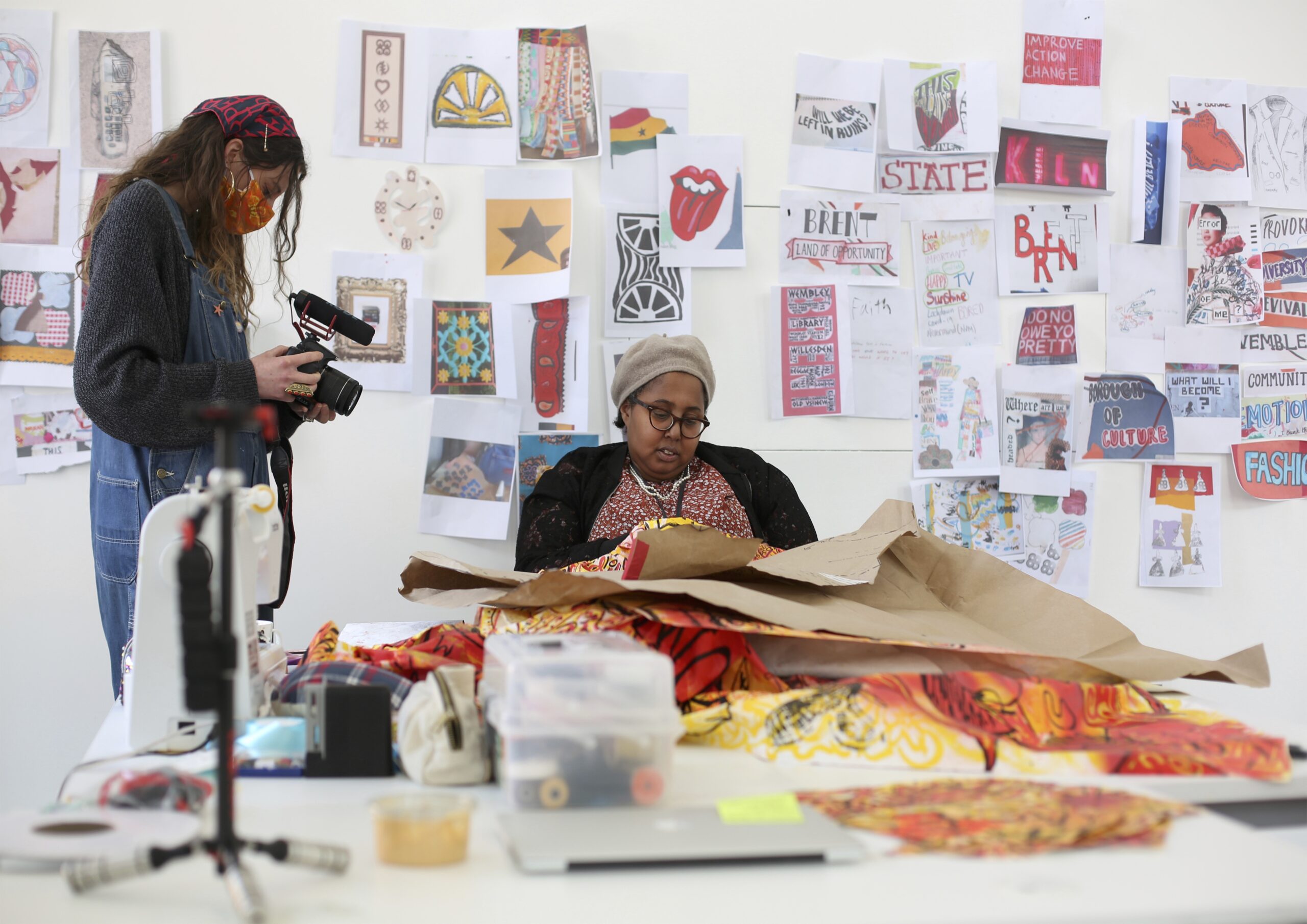 A woman stands with a video camera in an art gallery. She is filming a Black woman wearing glasses who is sat down and busy dressmaking. On the walls behind them are colourful works of art.