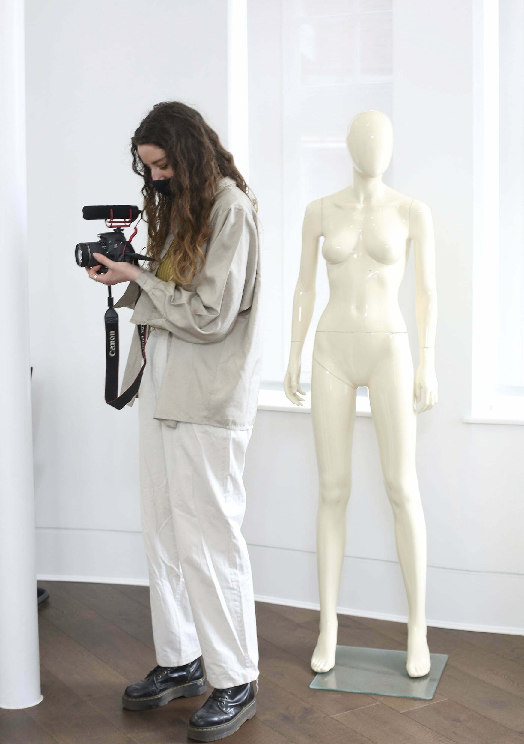 A woman dressed in a white sweatshirt and whte trousers looks through a film camera viewfinder in a white studio. A white undressed manequin is behind her.