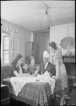 Black and white image of a mixed race Muslim family in the 1940s sat around a dining table in a typical British home.