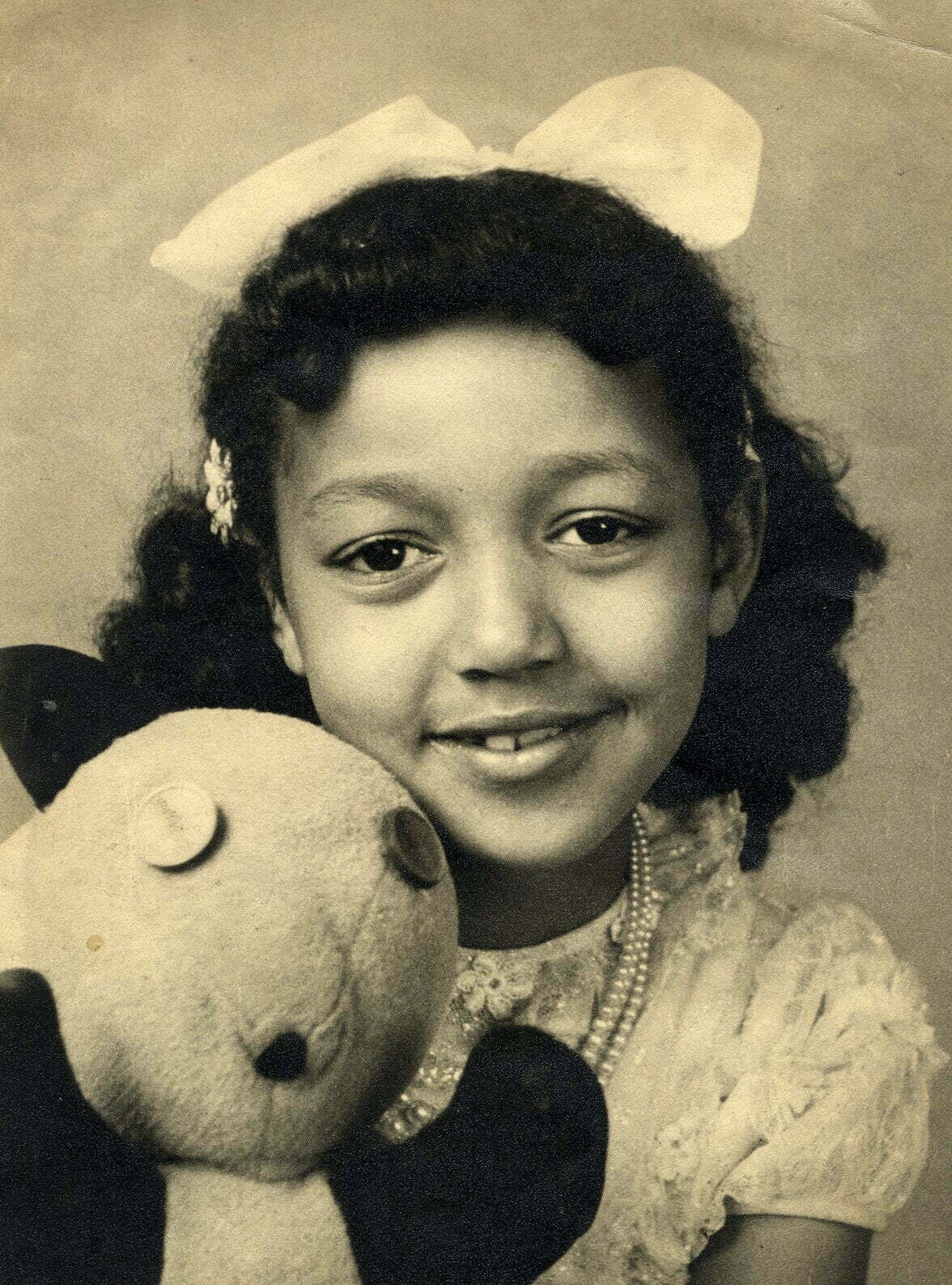 Black and white studio photograph of SuAndi as a young girl with a bow in her hair, holding a teddy and smiling at the camera.
